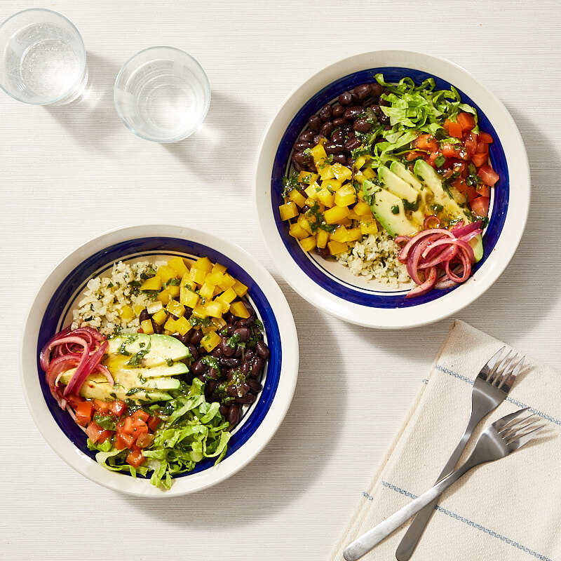 Two individual serving bowls filled with cauliflower rice and topped with black beans, chopped, tomatoes, shredded romaine, sliced avocado, chopped yellow pepper, and minced onion, drizzled with dressing