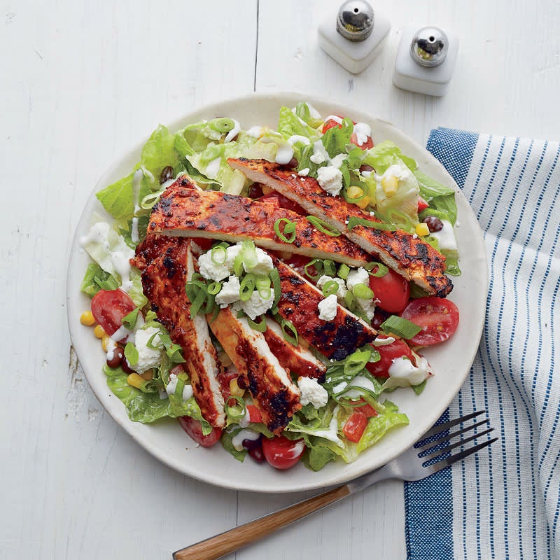 large white dinner plate topped with lettuce, tomatoes, corn, black beans, peppers, goat cheese, and sliced, grilled BBQ chicken, drizzled with ranch dressing