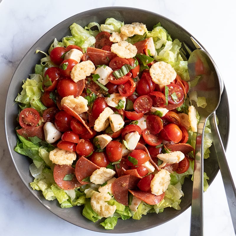 large gray serving bowl filled with lettuce, tomatoes, sliced pepperoni, and fresh mozzarella balls tossed with vinaigrette and topped with Parmesan crisps