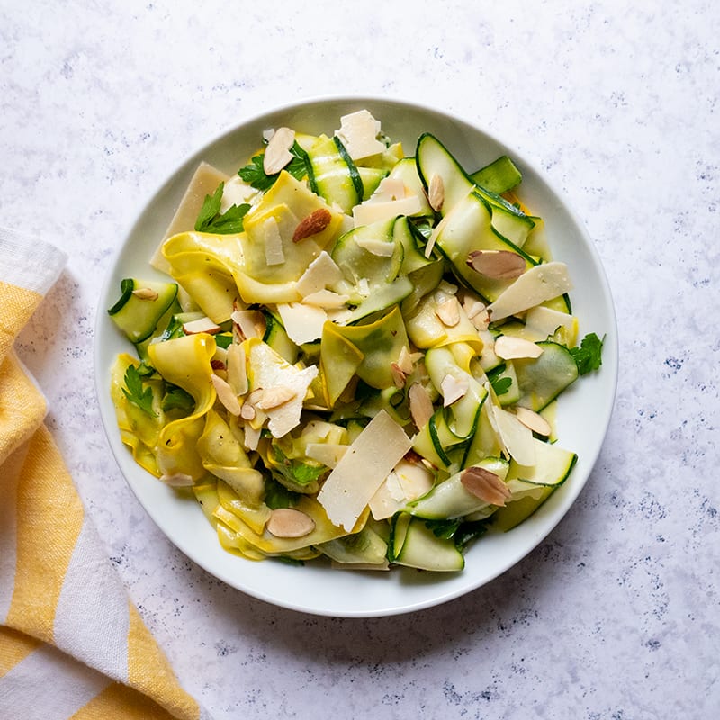 Round serving dish filled with vinaigrette-drizzled summer squash ribbons topped with parsley, almonds, and Parmesan cheese