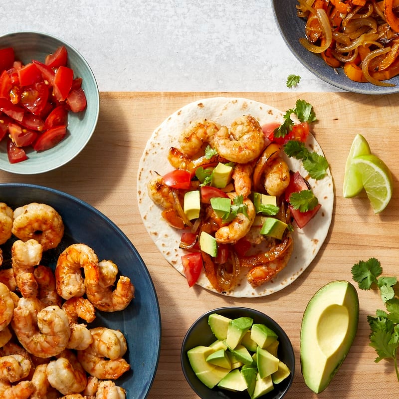 a wooden surface with individual bowls of cooked shrimp, cooked peppers and onions, chopped tomatoes, and chopped avocado with an already prepped fajita on the surface