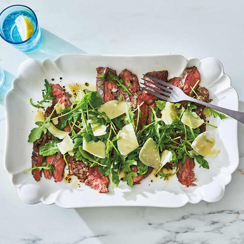 Rectangular white platter topped with arugula salad, thinly sliced grilled steak, and shaved Parmesan cheese