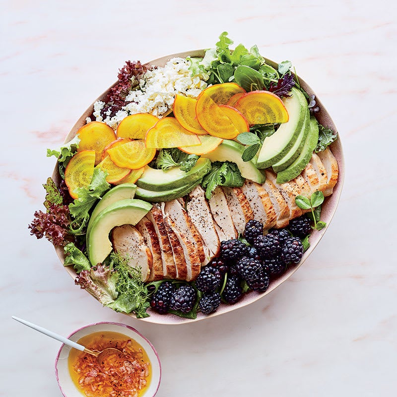 Large oval platter filled with lettuce and topped with rows of blackberries, sliced chicken, sliced avocado, sliced golden beets, and feta cheese, served with vinaigrette on the side