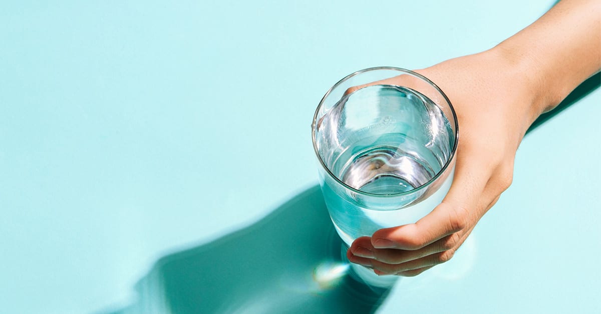 A person's hand holding a clear glass of water, with a shadow on a turquoise surface.
