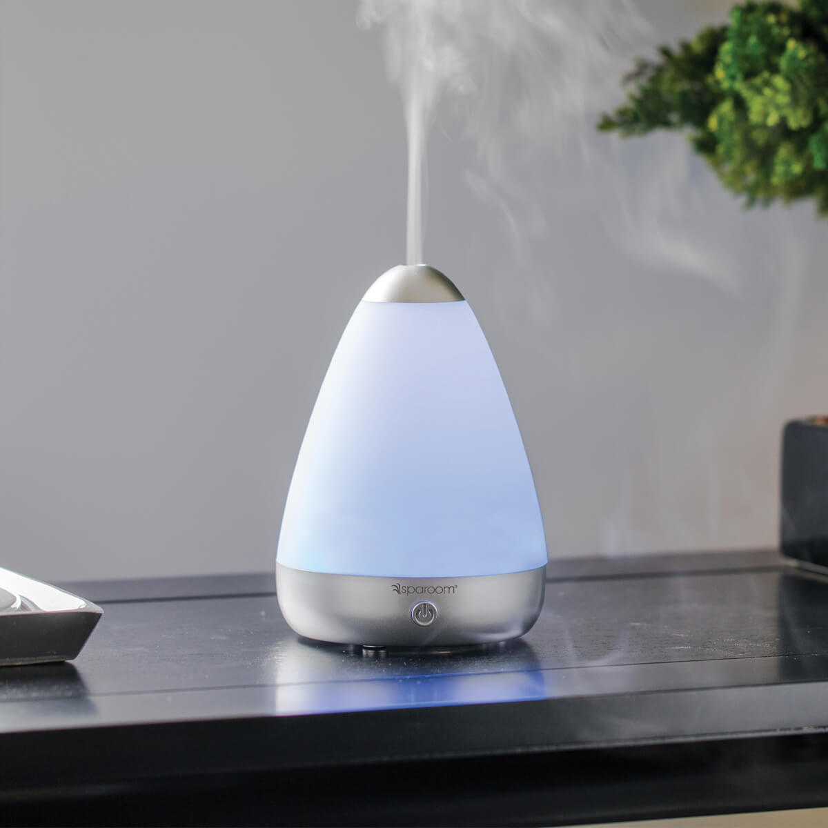 A white pear-shaped device sits on a black counter. It is streaming a mist into the air, which is floating toward the small plant sitting next to it.