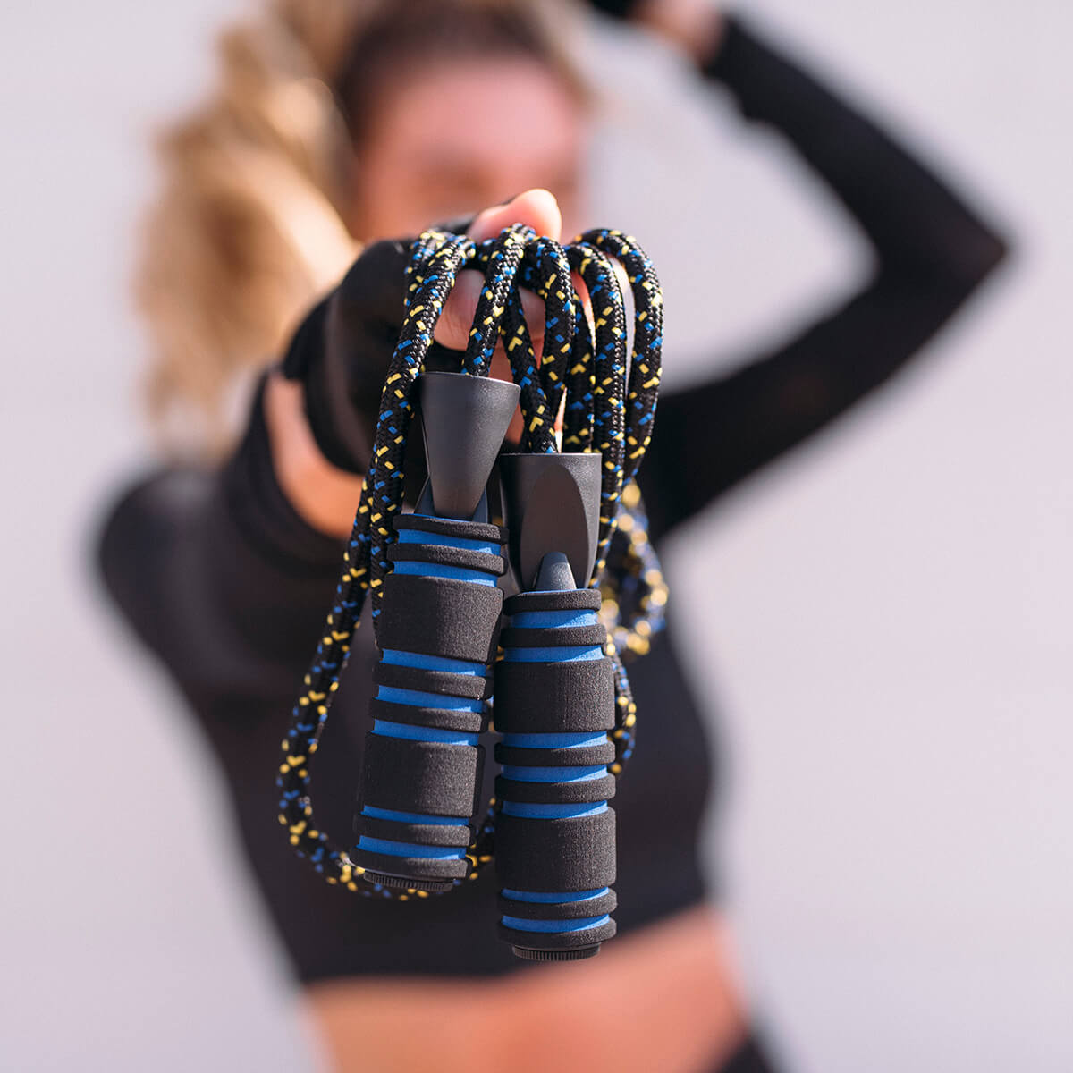 A blonde woman wearing black and showing her midriff hands a jump rope toward the camera. The black ripe is connected to a pair of black handles with seven blue stripes.