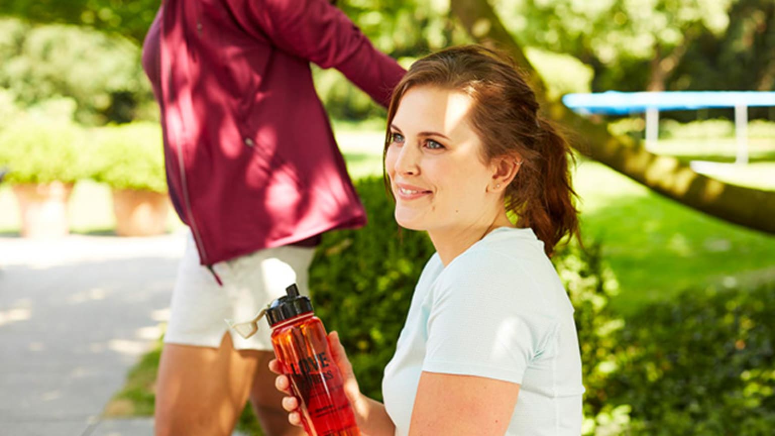 Junge Frau in weißem Sportshirt sitzt mit Trinkflasche auf einer Parkbank.