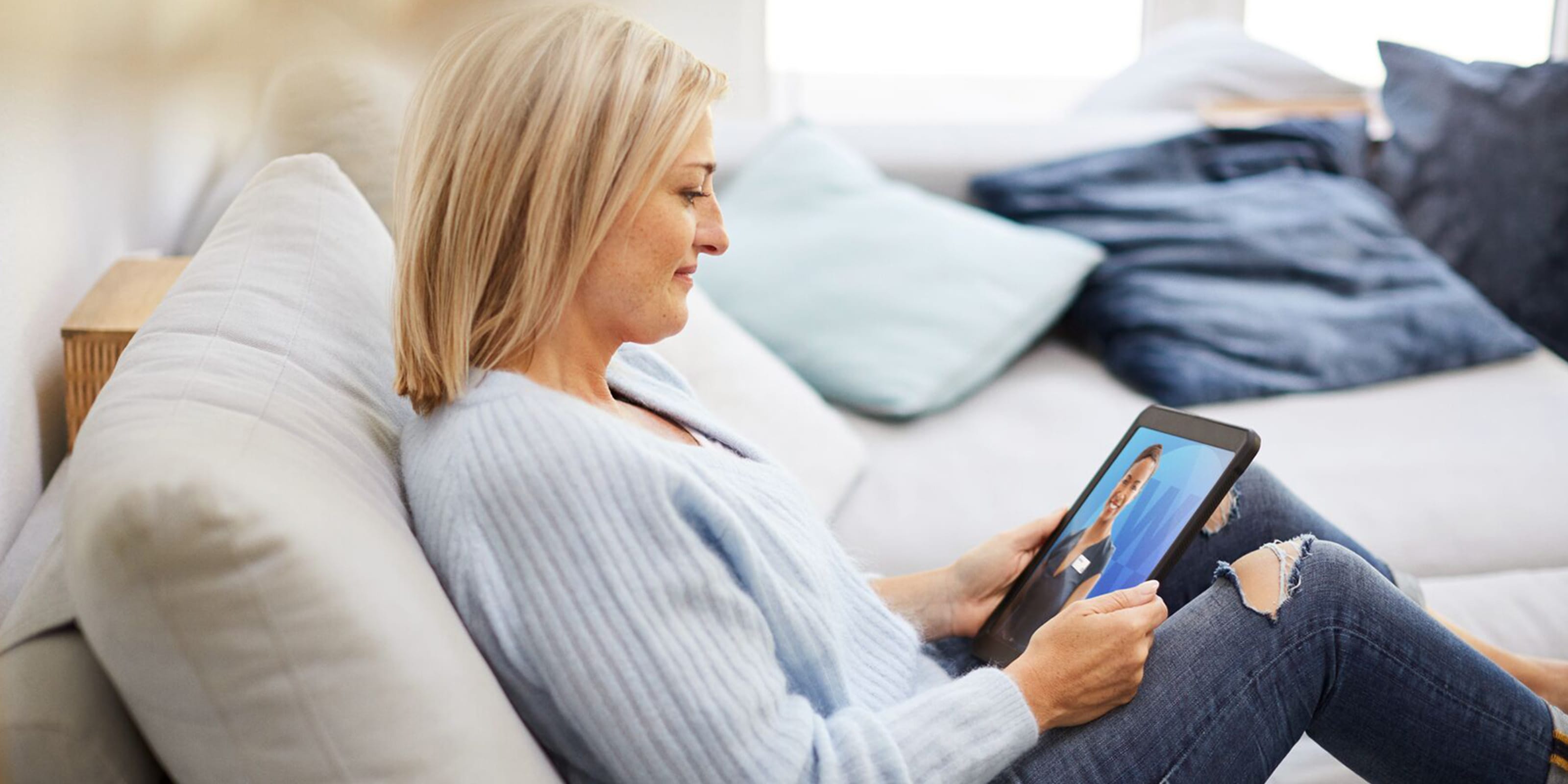 Une femme blonde en pull bleu clair est assise sur un canapé avec une tablette à la main.