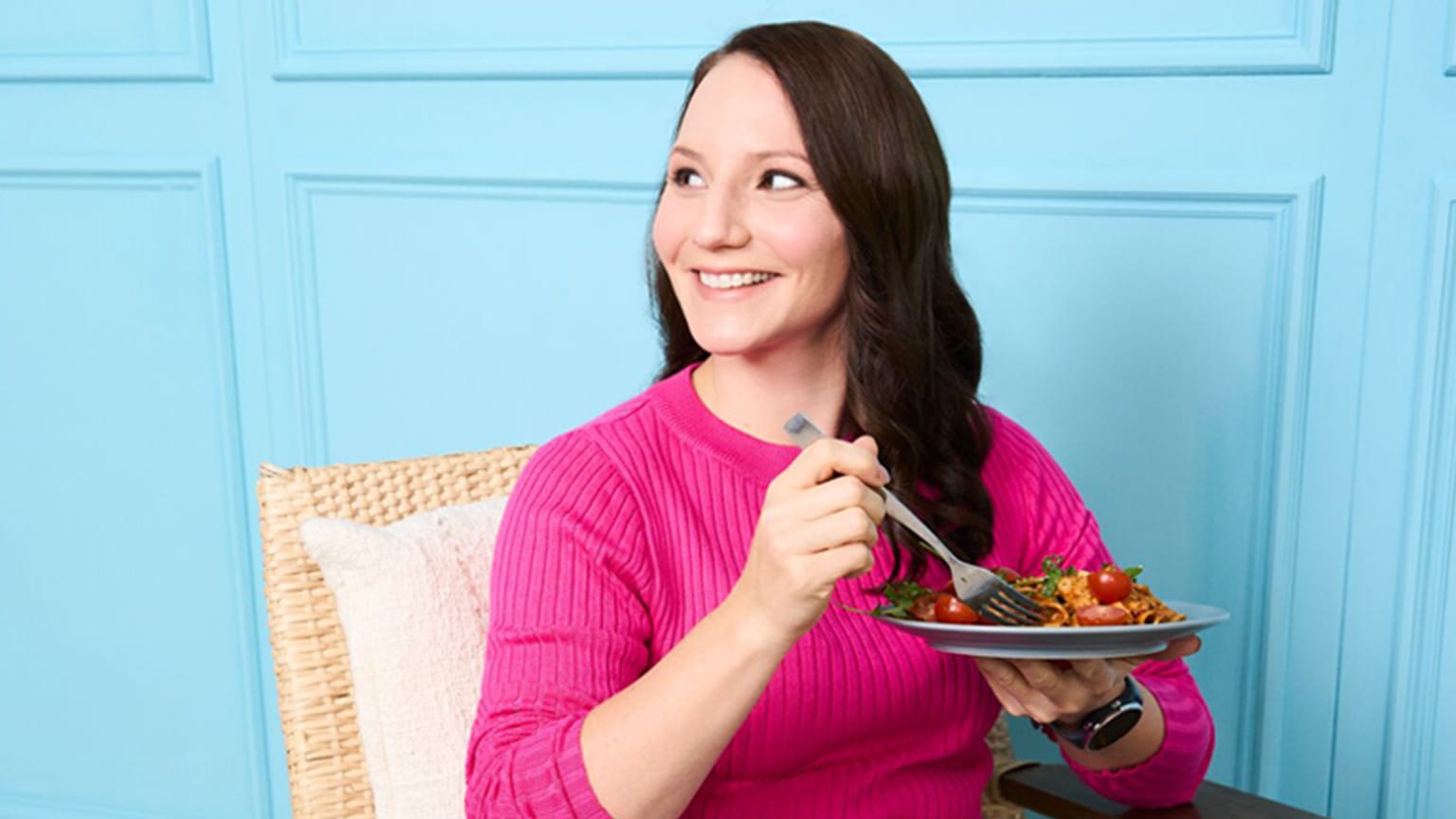 Junge Frau mit braunen Haaren und pinkem Pullover sitzt mit Pastateller vor blauer Wand.