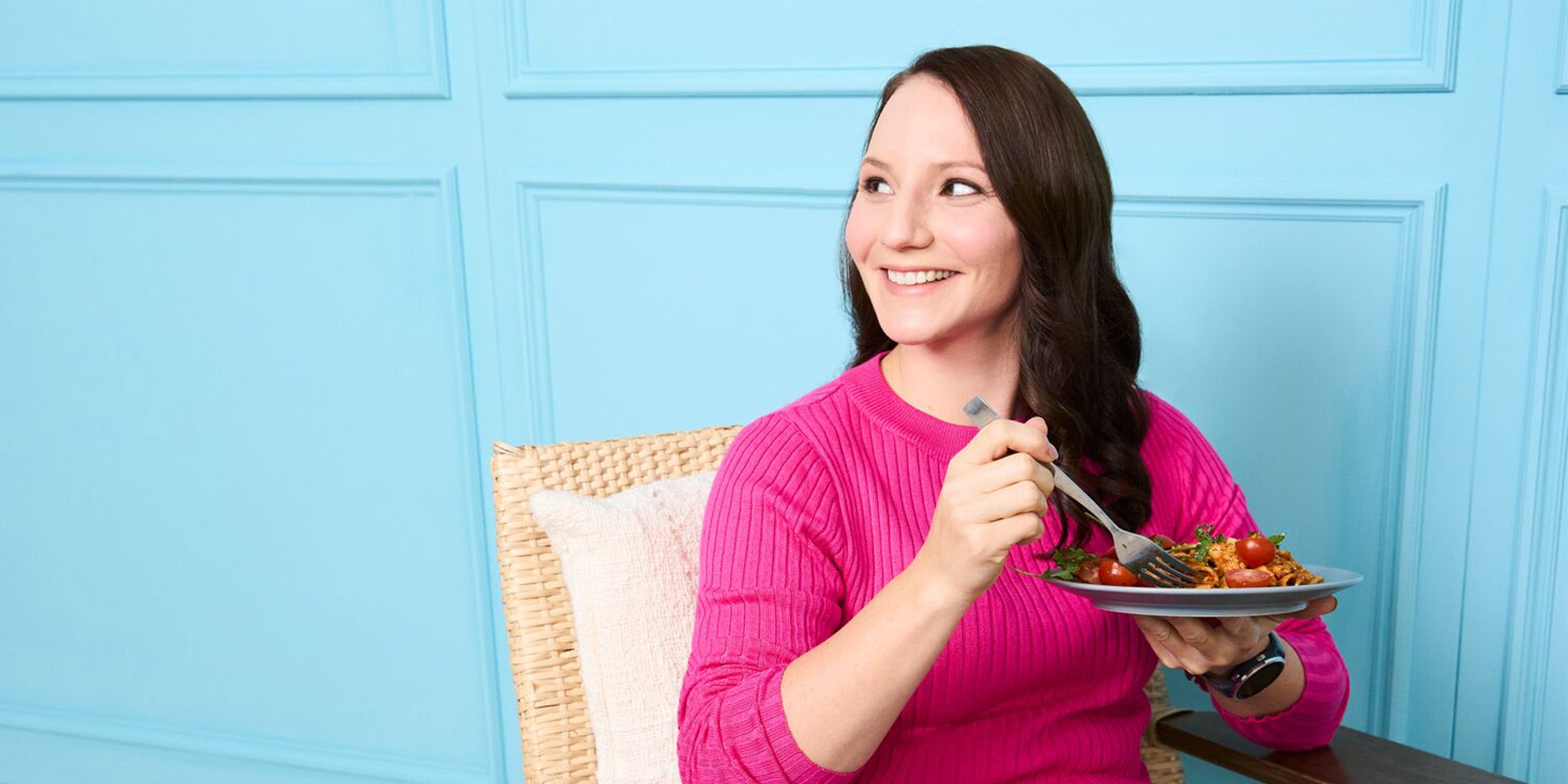 Junge Frau mit braunen Haaren und pinkem Pullover sitzt mit Pastateller vor blauer Wand.