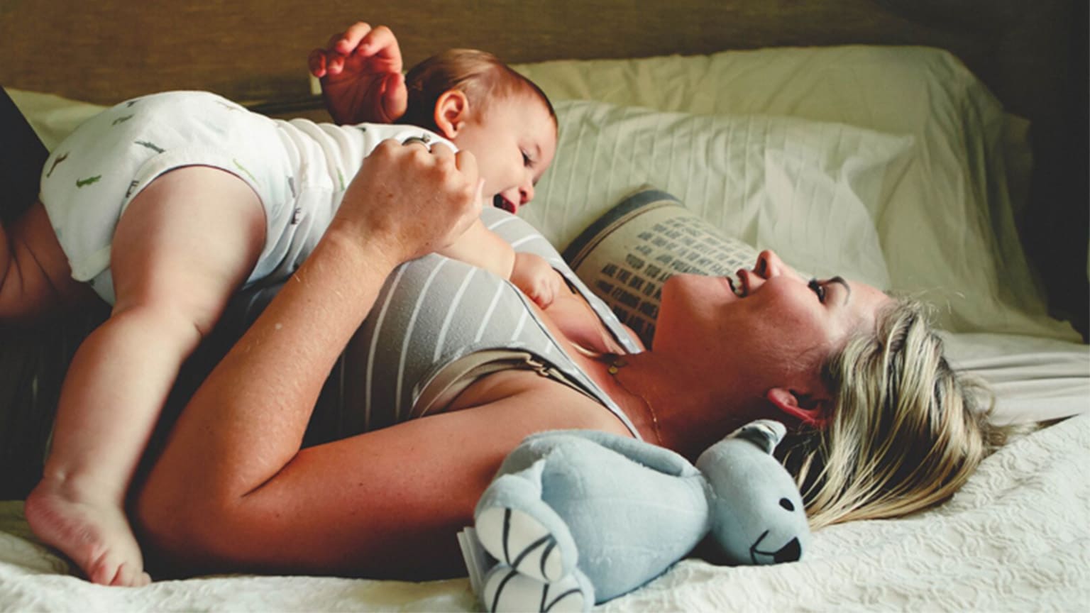 Une femme aux cheveux blonds est allongée sur un lit avec un bébé et rit.