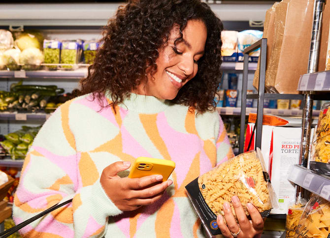 Woman in a grocery store holding a bag of pasta in one hand and her iphone in the other, while smiling.