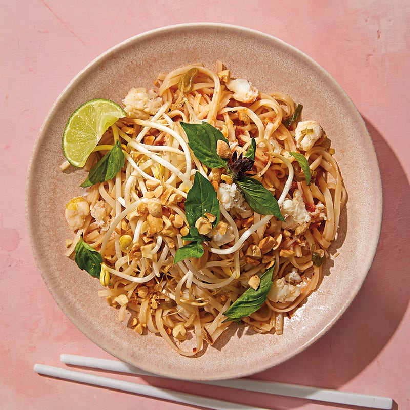 pink bowl filled with Crab Pad Thai, garnished with a fresh lime wedge, chopped peanuts, and torn fresh basil