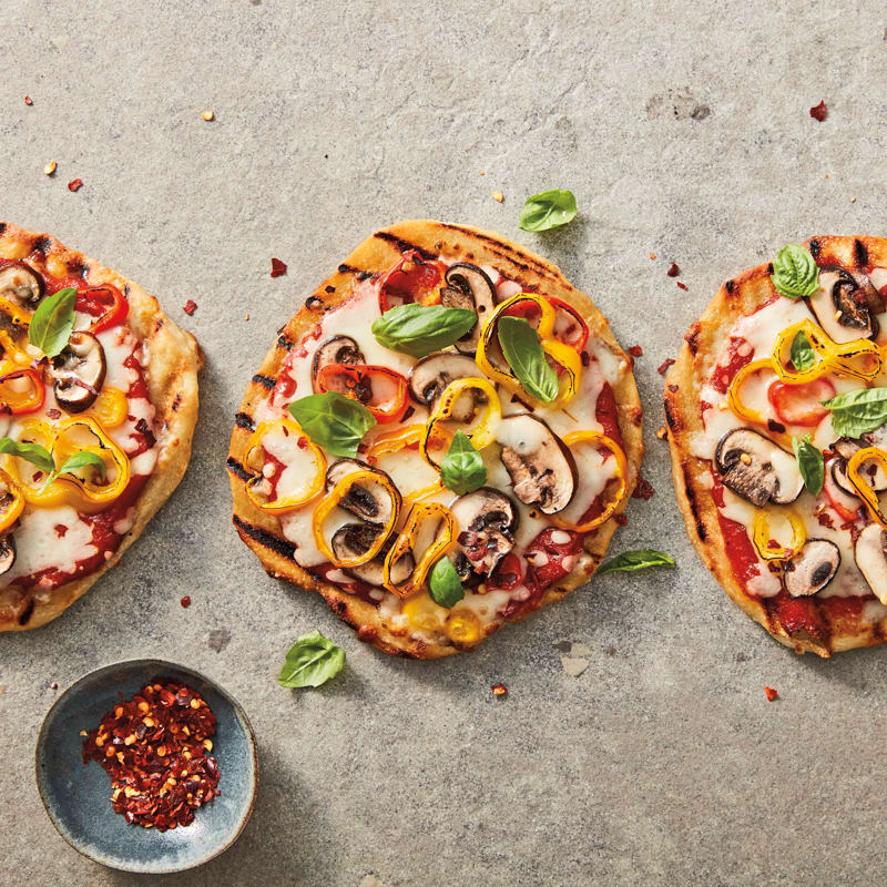 Three individual grilled mushroom and pepper pizzas on a surface, served with a small bowl of red pepper flakes