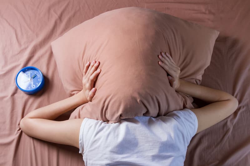 Mulher deitada na cama cobrindo a cabeça com um travesseiro e um relógio azul ao lado dela