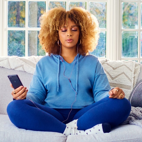 Junge Frau mit Afrofrisur sitzt meditierend in gemütlicher Kleidung auf dem Sofa 