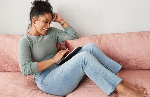Junge Frau in Jeans sitzt auf einem rosa Sofa und schaut auf ihre Tablet
