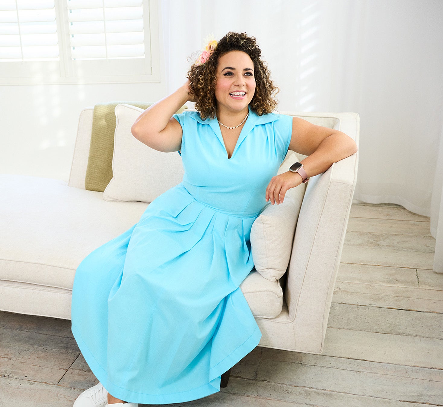Junge Frau mit lockigen Haaren sitzt in hellblauem Kleid auf beigem Sofa