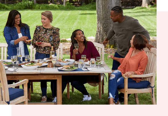 5 people are sitting and standing around a wooden table in a backyard on a sunny day. On the table are plates of food and glasses of white wine.