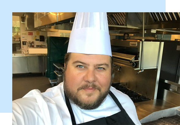 Rodney Bowers stands in a kitchen setting, smiling wearing while an apron and chefs hat